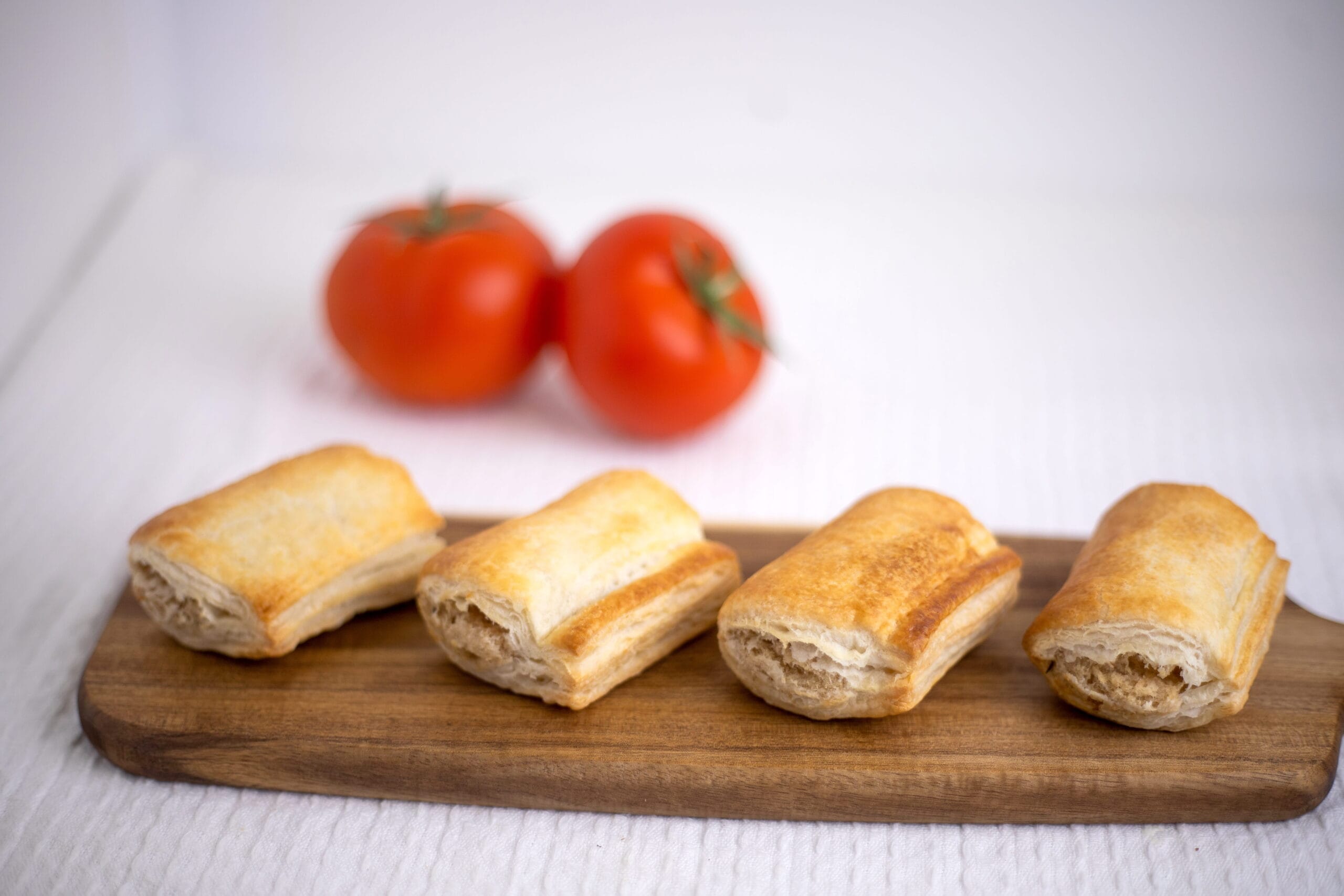 Golden puff pastries on a wooden board with blurred red tomatoes in the background.
