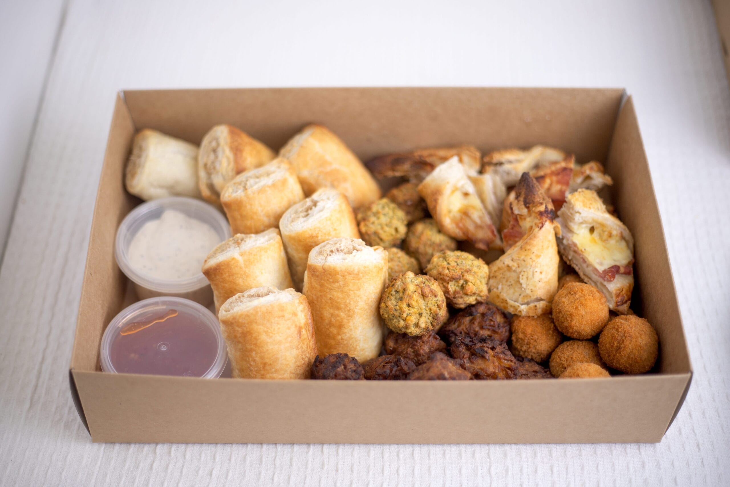 Box of assorted bite-sized pastries and fried snacks with dipping sauces in plastic cups
