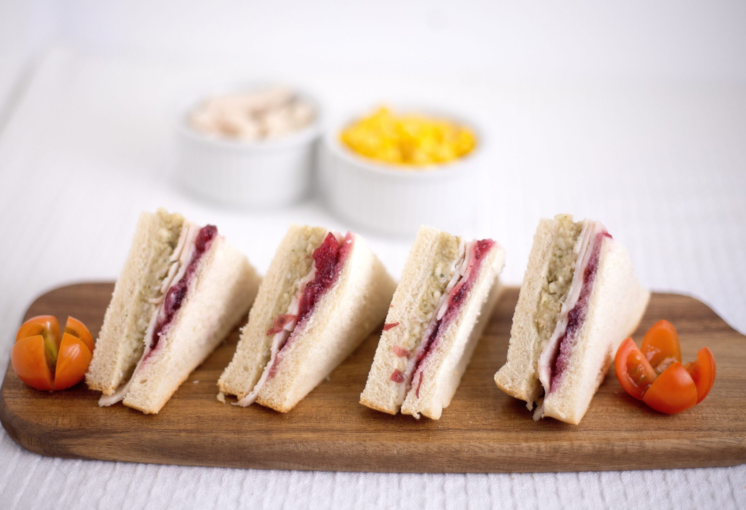 Crustless sandwich wedges with pale filling and red jam on a wooden board, with small orange peppers nearby.