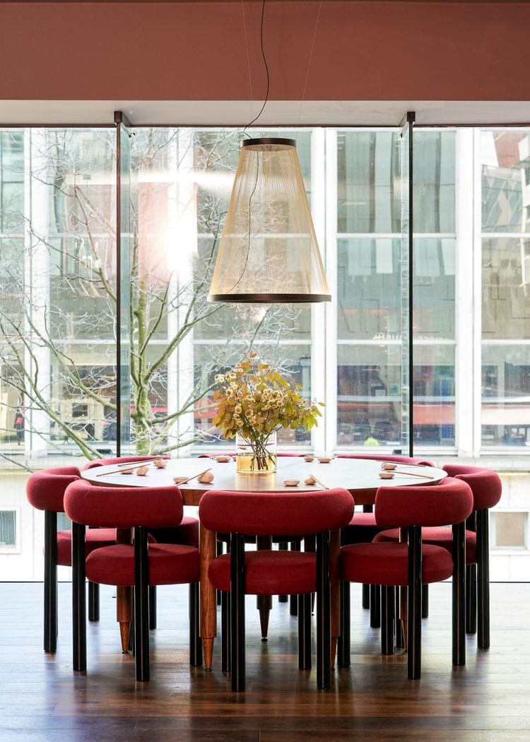 Round wooden dining table with red upholstered chairs around it, a vase of flowers center, under a large pendant light in a glass-walled room.