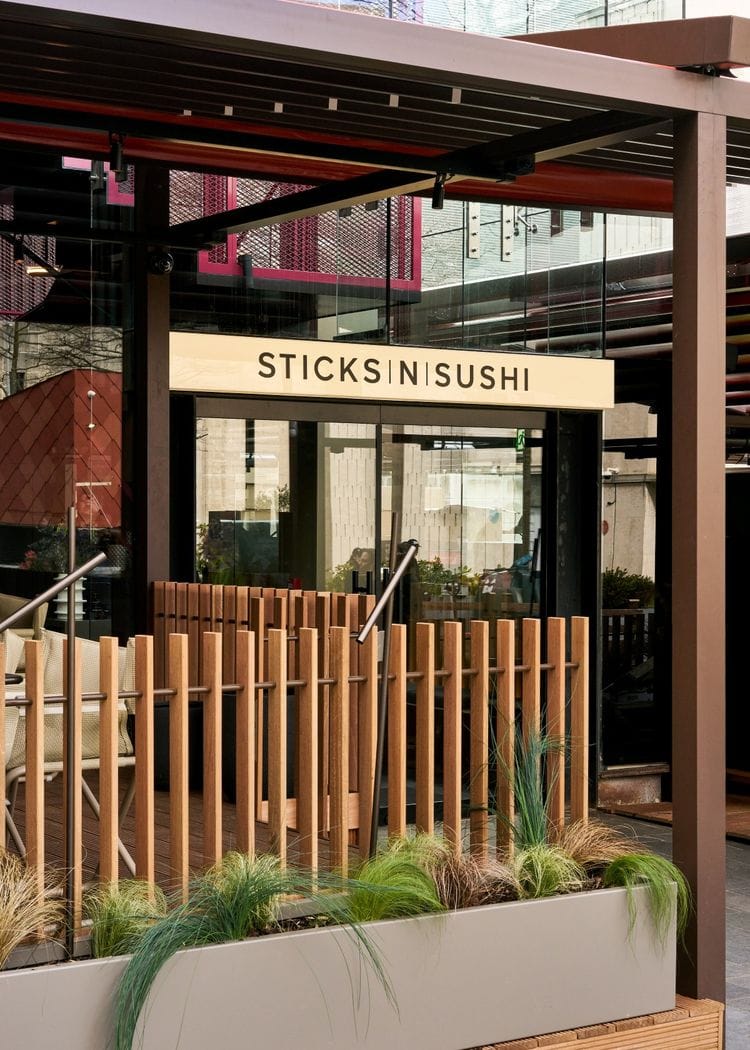 Exterior of a sushi restaurant called STICKS|N|SUSHI with a wooden slat fence and outdoor seating