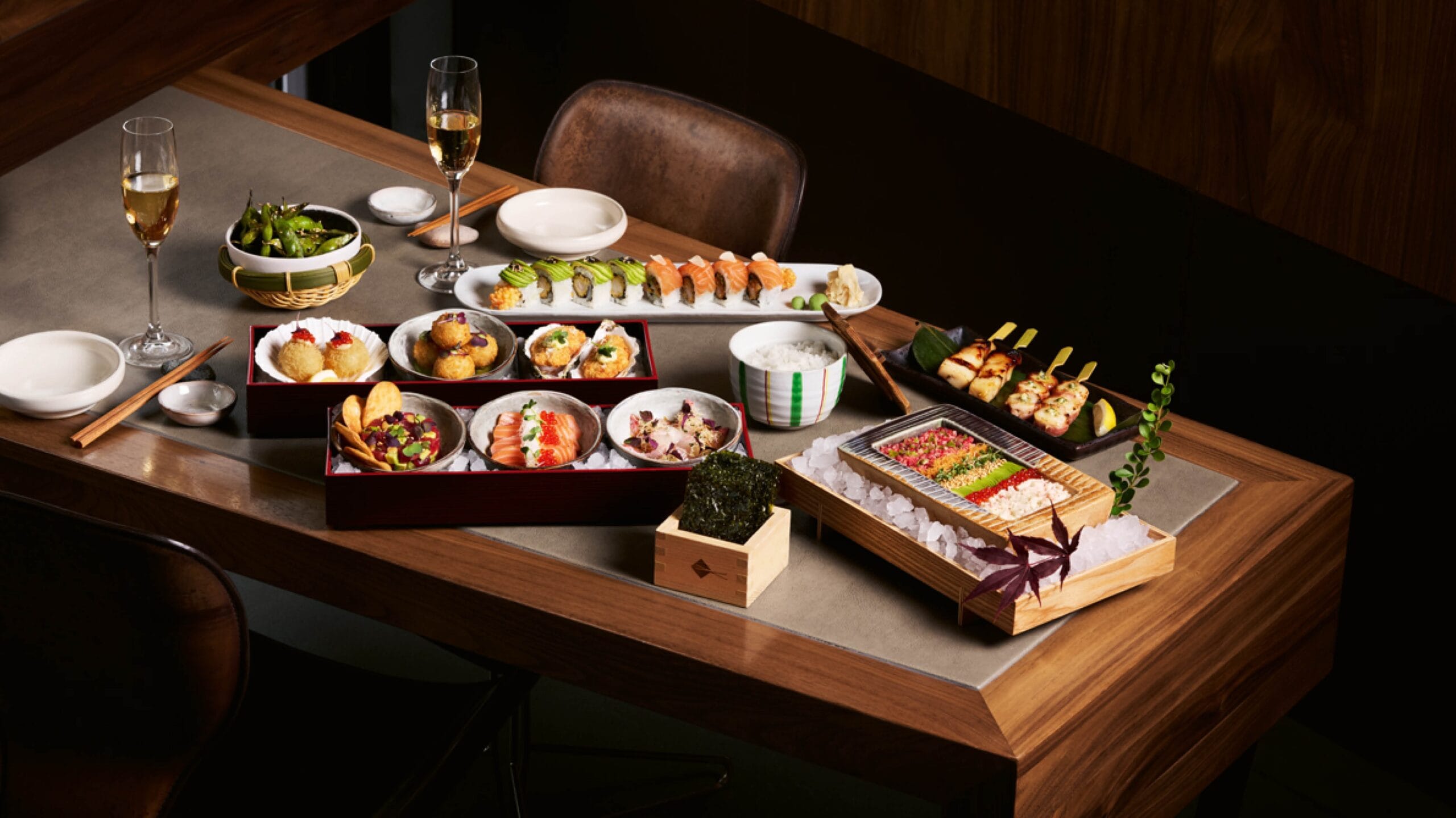 Assorted sushi and Japanese dishes arranged on a dining table with two wine glasses in the background.